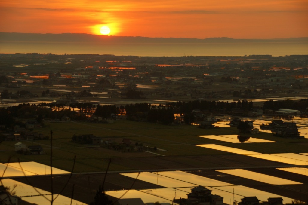 夕方の田園風景を撮影した写真。高い場所から撮影されており、広大な田園風景は夕焼けの影響でオレンジ色に染められている。