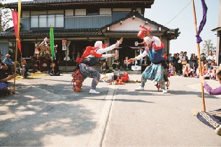 手踊りを行っている写真。住宅前の道路で二人の大人が踊っており、周りには踊りを見る大勢の人がいる。