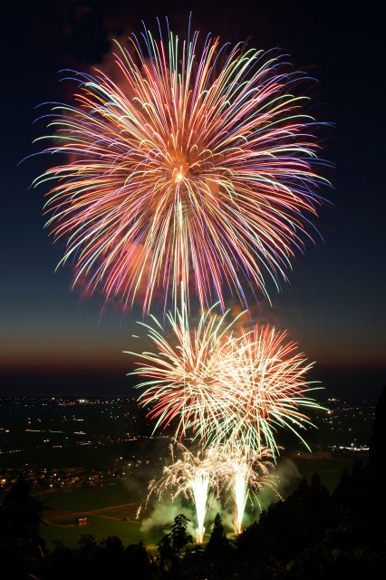 打ち上げ花火の写真。夜空の下、高い場所から撮影した写真で、爆発後に散った花火は綺麗な光の線を描いている。