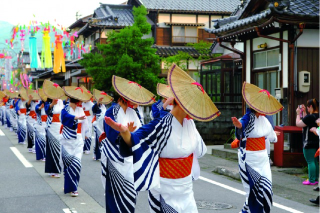 祭りの写真。車道の真ん中で着物をきた多人数の大人が列を作り、踊りを踊っている写真。