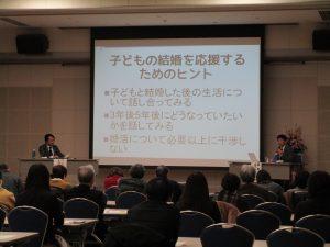 たくさんの参加者が大きなスクリーンを使って話す講師のセミナーを聞いている様子の写真
