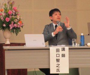 右手にマイクを持ち、座りながら話をしている田口智之氏の写真