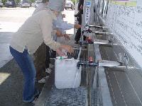 屋外の蛇口が並ぶ給水場で、町民が飲料水用ポリタンク片手に水を汲んでいる写真