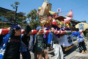 青空の元、大きな鯛や小槌などが飾られたお神輿の二天棒を担ぐ法被姿の男たちの写真