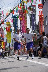旧北陸街道の面影を残す通りが七夕飾りで彩られるマラソン大会で半袖短パンの学生二人がたすきを渡す瞬間の写真