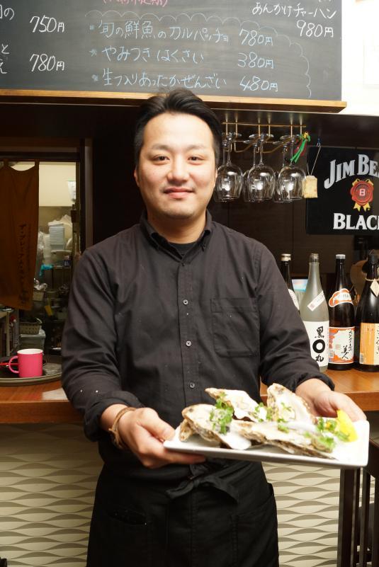 黒シャツに黒エプロン姿の料理人が、お店のカウンター前で牡蠣料理を手に持っている写真