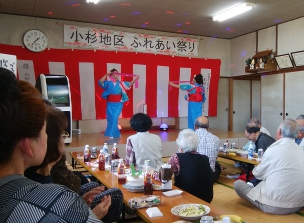 小杉区ふれあい祭りを撮影した写真。屋内で宴会が行われており、奥にはステージで盆踊りを披露している女性2人、手前には食事しながら披露されている盆踊りを見て楽しでいる多くの人々が写っている。