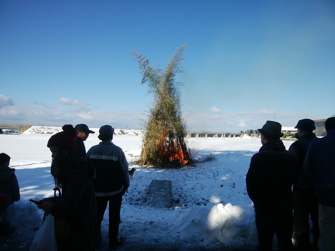 雪の上で左義長が行われている写真。写真の中央には大きな木が2本燃やされており、火の周りには左義長を見守る人々がいる。