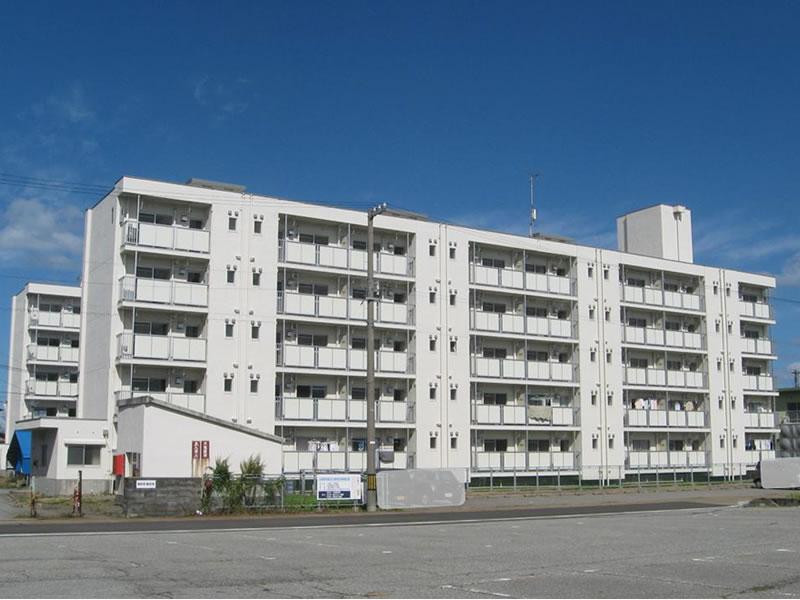 白い建物の写真。建物は5階建てで手前と奥に2棟並んでいる。また建物の手前には広い駐車場もある。