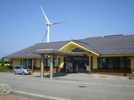 青空の下に瓦屋根と黄色い外壁が目立つ平屋建ての入善浄化センター管理棟と大きな白い風車の写真
