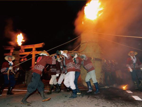 タイマツ祭りの最中の写真。大きな藁には神輿のような持ち手が付けられており、この大きな藁を複数の男性が神輿を担ぐように背負っている。また藁のてっぺんには大きな炎が燃えている。