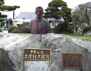 巨大な庭石と松の木が映える庭園の岩の上に、長い白髭に面長で丸眼鏡をかけた初老の紳士のバストアップの赤茶けた銅像が設置されている写真