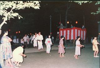 夜、赤と白の幕を張った櫓の周りを囲み盆踊りを楽しむ人々の写真