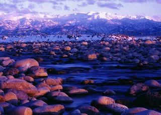 夕暮れ、薄紅色に染まる雪化粧の北アルプスと岩々の間を流れる川の流れ