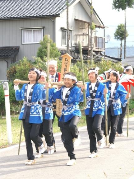 竹に吊るされた米俵を担いだ笑顔の女子小学生たちが練り歩いている様子の写真
