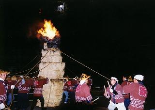 火のついた藁でできた巨大松を法被にヘルメット姿の人々が夜空のもと練り歩く写真