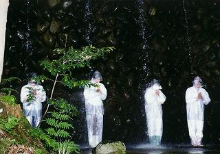 白い装束に身を包んだ4人の人が黒い岩肌を背に滝に打たれている写真