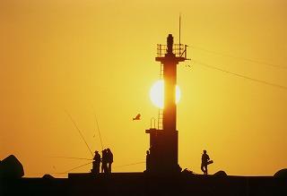 夕日の中、堤防で釣りを楽しむ人々と堤防上の灯台のシルエット。夕日が灯台の後ろにかかり幻想的な雰囲気を見せている写真