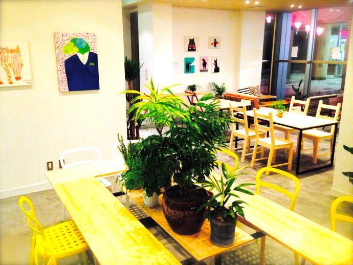 お洒落な黄色のイスと観葉植を中央並べたテーブル、壁には小さな絵画が並べてある明るいカフェテラスの写真