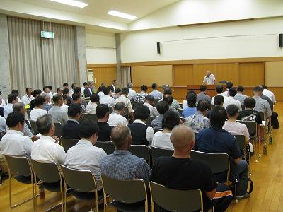 公民館のホールで行われている「まちづくり懇談会」にて、檀上でしゃべる人と、沢山並べたパイプ椅子に座って話を聞く町民達の写真